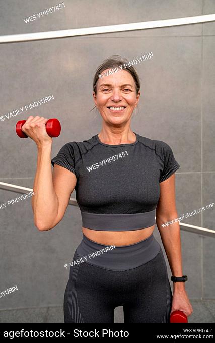 Smiling woman exercising with dumbbells in front of wall