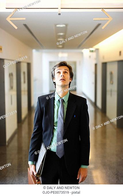 Confused businessman in corridor looking up at arrows pointing opposite directions