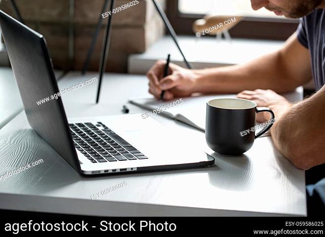 Man with a beard is using a laptop on the blurry background of the window. Laptop is on the white wooden table with a cup near it