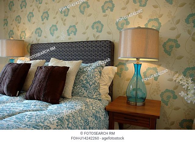 Lighted lamps with blue glass bases on either side of bed with blue and white duvet in modern bedroom with blue stylised floral patterned wallpaper