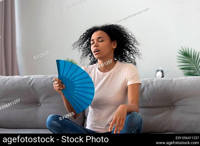 Tired African American woman suffering from heat stroke at home, waving blue fan, enjoying fresh air, sitting on couch, sweaty young female cooling in hot...