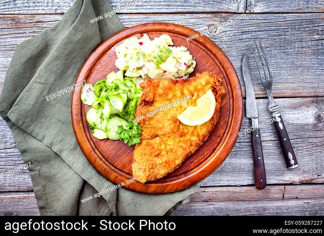 Traditional deep-fried schnitzel with potato and cucumber salad offered as top view on a rustic wooden board with copy space