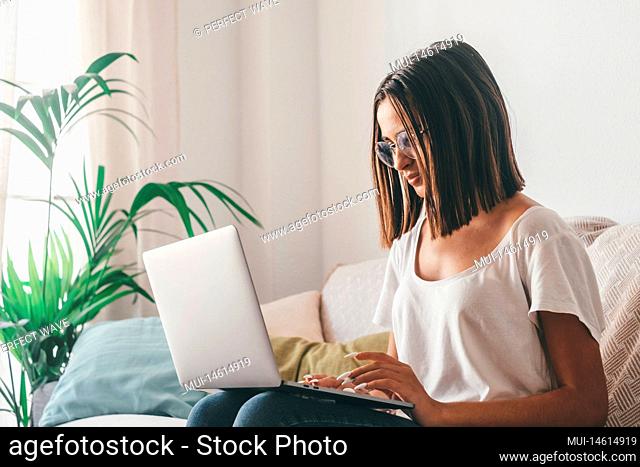Happy young woman in eyeglasses typing using laptop while sitting on sofa in the living room of her house. Beautiful lady spending leisure time using laptop