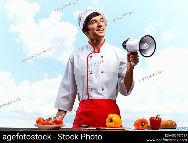 Young chef standing with megaphone in hand. Emotional caucasian chef in white hat and red apron on blue sky background. Restaurant advertisement