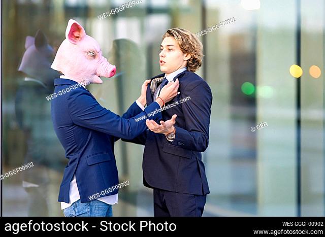 Businessman in pig mask holding collar of male colleague by glass wall