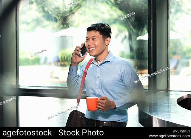 Portrait of a handsome and cheerful Asian young man talking on mobile phone, while holding a cup of coffee in the modern reception area of a business building