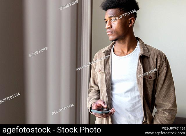 Thoughtful young man with smart phone looking out of window at home