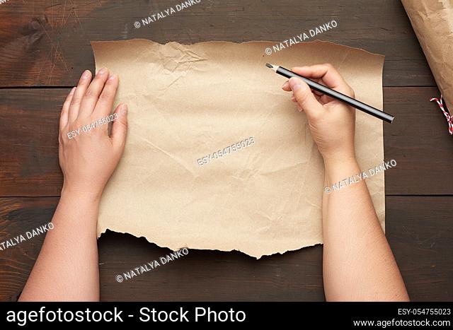 empty torn sheet of brown craft paper and two hands with a black wooden pencil, wooden table, top view, place for an inscription