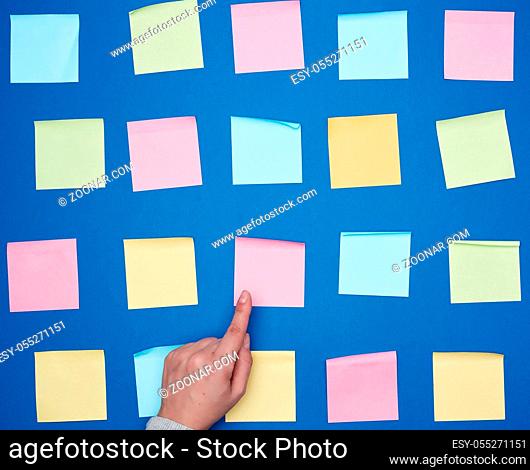 female hand and a lot of empty paper multi-colored square stickers on a blue background, concept of choosing a strategy with the specified finger