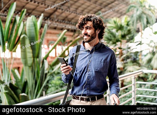Smiling businessman wearing in-ear headphones standing with smart phone by plants