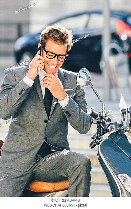 Smiling businessman sitting on motor scooter talking on cell phone