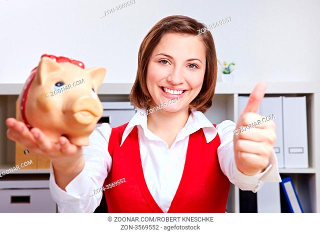 Lachende Geschäftsfrau mit Sparschwein im Büro hält ihren Daumen hoch