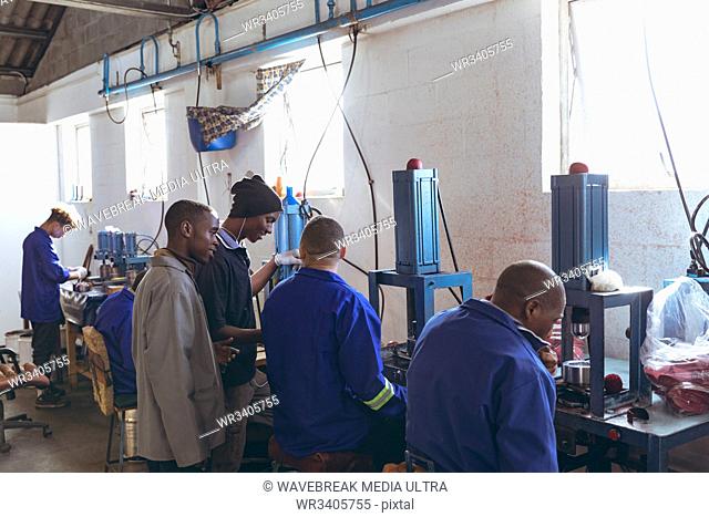 Side view of a diverse group of young men working at a cricket ball factory talking together while they operate machinery in the production line