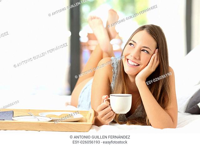 Happy girl having breakfast holding a coffee cup lying on the bed and looking sideways