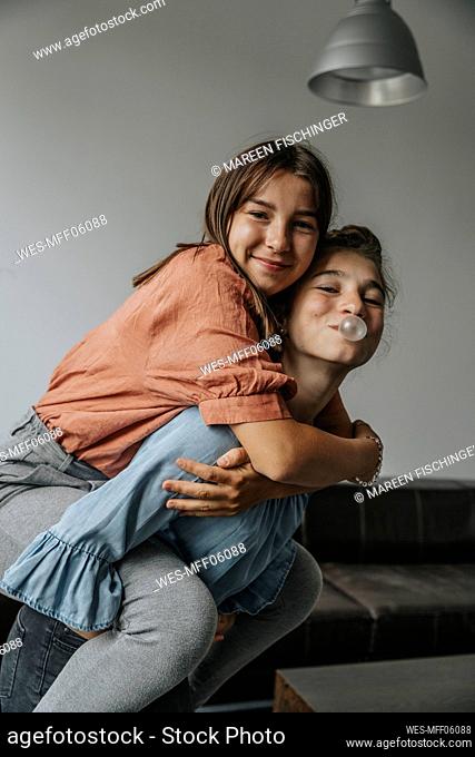 Playful girl blowing bubble gum while piggybacking friend against wall at home
