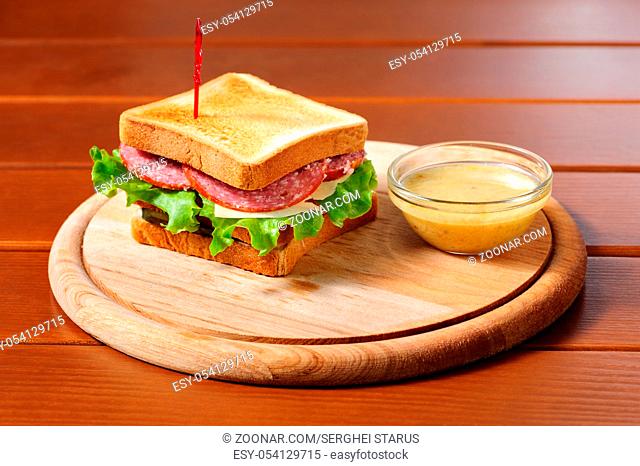 toast sandvich with salami, cheese, meat, pickles and lettuce at round wooden plate and honey mustard sauce