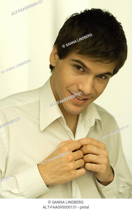 Young man buttoning shirt, smiling, portrait