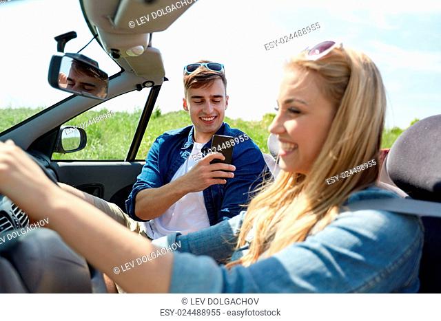 leisure, road trip, travel, summer holidays and people concept - happy couple driving in cabriolet car and taking picture by smartphone