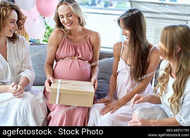 Smiling pregnant woman opening gift box by friends on sofa