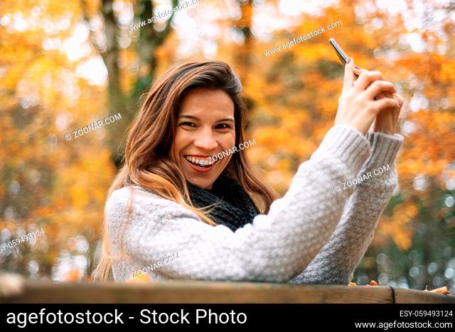 Woman laughing holding her mobile phone and looking at camera in Autumn city park
