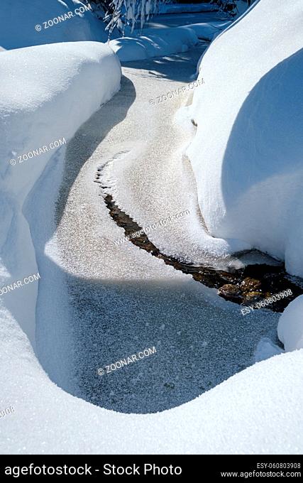 Small frozen stream with snowdrifts on shores in mountain snowy winter forest