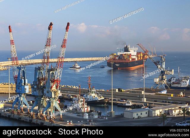 Dock with four-link cargo loading cranes and docked tugboats plus Dimitris Y container ship heading out to sea, Ashdod Port, Israel