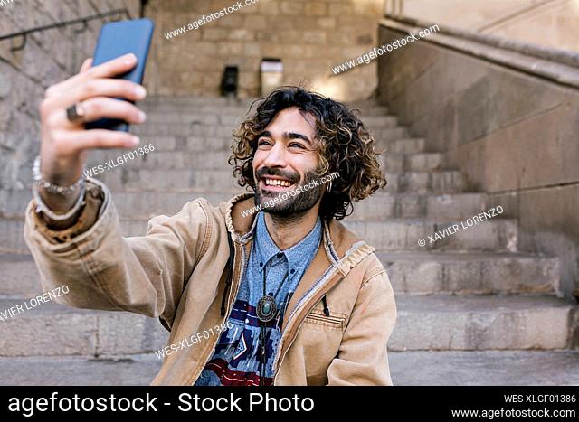 Happy hipster man taking selfie through mobile phone while sitting on steps