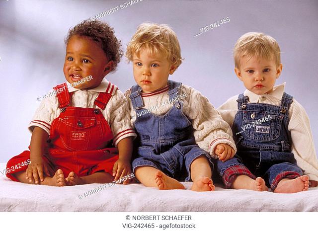 portrait, full-figure, indoor, group of 3 1-year-old children, 1 black and 2 blond boys wearing white pillover and jumpers sit on a terry towel  - GERMANY