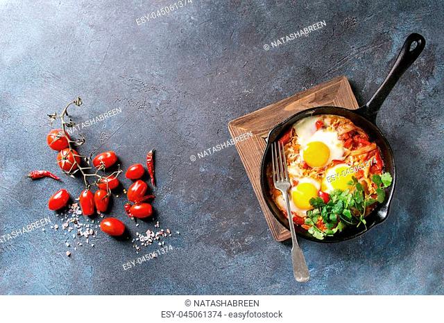 Traditional Israeli Cuisine dishes Shakshuka. Fried egg with vegetables tomatoes and paprika in cast-iron pan on wooden board with coriander herbs over blue...