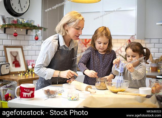 Blond woman wearing blue apron and two girls standing in kitchen, baking Christmas cookies