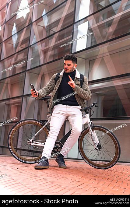 Male professional gesturing while taking selfie leaning on bicycle by office building
