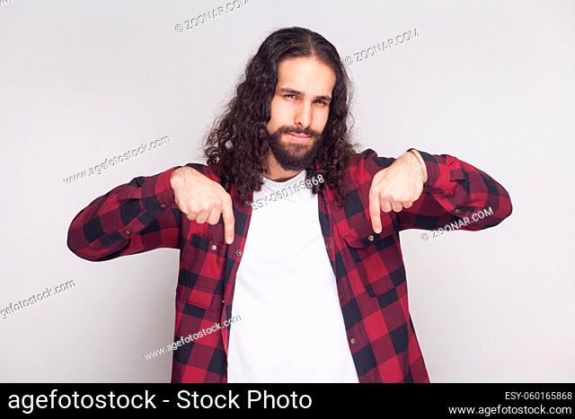 Here and right now. serious handsome man with beard, black long curly hair in casual checkered red shirt standing, looking at camera and pointing down