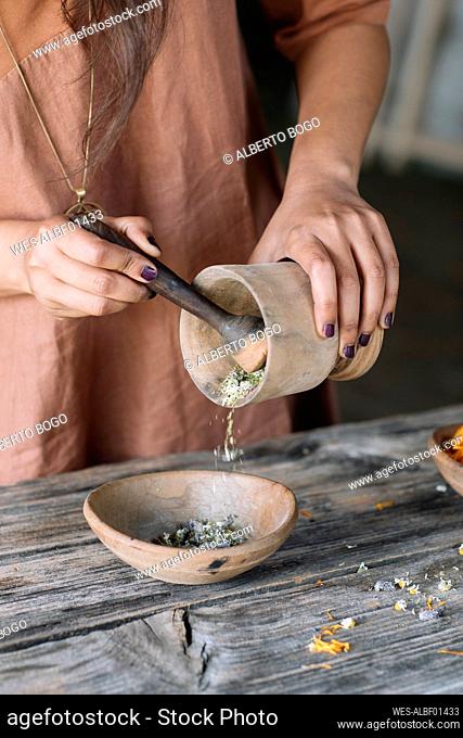 Midsection of woman pouring crushed flowers and herbs from mortar in earthenware bowl on wooden table