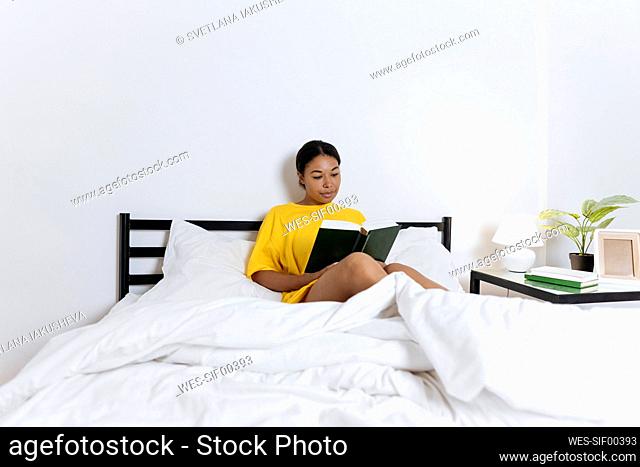 Woman in yellow t-shirt sitting on bed reading a book