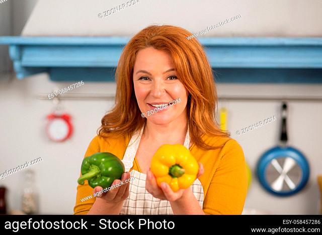 Close up view of red haired housewife holding green and yellow peppers. Bright vegetables in hands of mid haired woman
