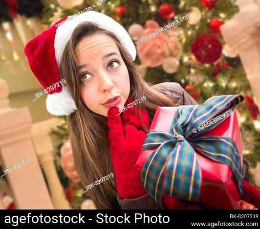 Thinking girl wearing A christmas santa hat with bow wrapped gift in front of decorated tree