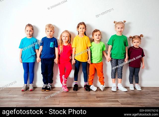 Lovely boys and girls in colorful sportswear posing in front of wall, holding hands. Team of preschoolers standing in a row, looking at the camera