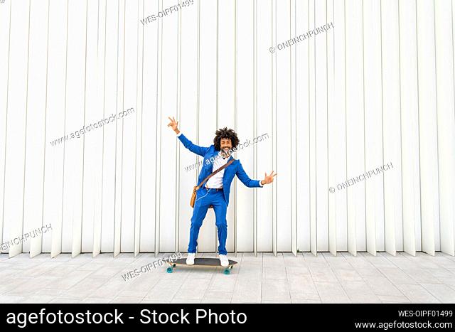 Playful businessman standing with arms outstretched on skateboard in front of wall