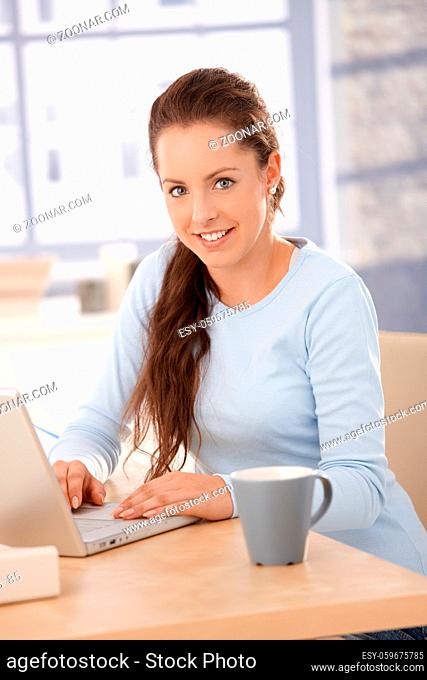 Attractive young female browsing internet at home, sitting at desk, using laptop, smiling