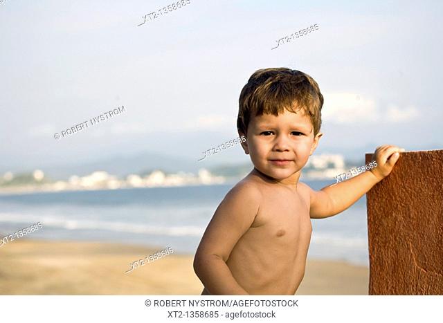 A young toddler stands against a wall beside the ocean and beach in Mexico