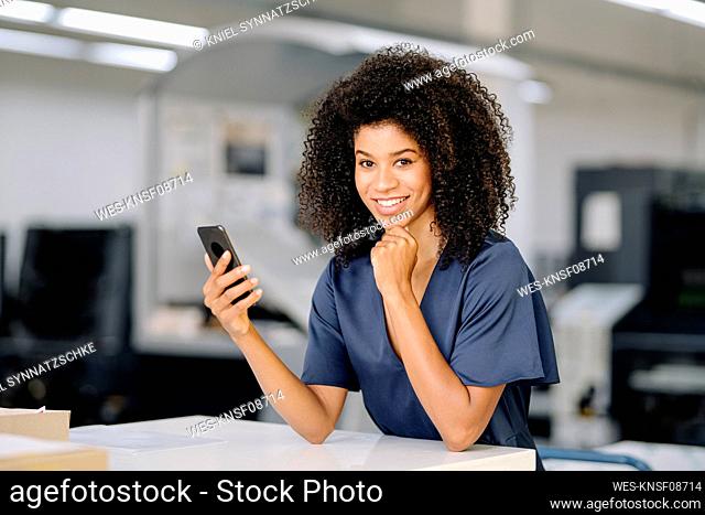 Smiling businesswoman with hand on chin holding smart phone while leaning on table at industry