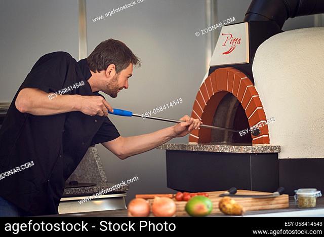 Middle-aged chef baking or flame grilling an Italian pizza in a modern stone oven at a pizzeria