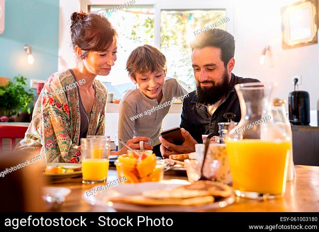Family breakfast, Dad shares a video on a phone to his son. Mom looks over her son's shoulder as the morning sun enters through the window