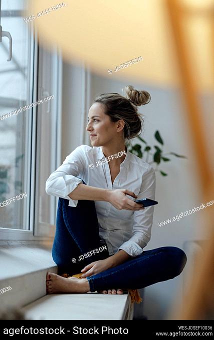 Smiling woman holding smart phone sitting on widow sill at home