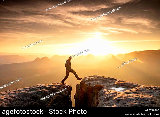 Strong mountain climber hiking and jumping over the summit ridge of a peaks at sunset. Man takes leap of faith off of rock outcropping