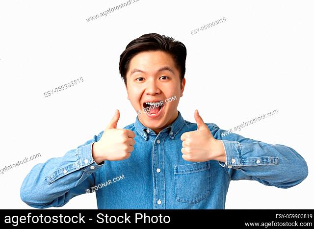 Close-up of successful winning, happy asian man with teeth braces smiling broadly and showing thumbs-up in approval, praising great work, saying well done