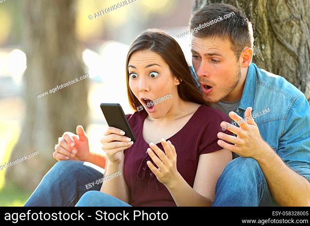 Surprised couple finding amazing content on line in a smart phone sitting in a park