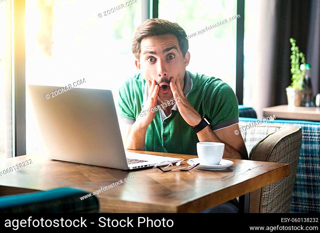 It's unbelievable. Young shocked businessman in green t-shirt sitting and working on laptop, looking at camera with surprised face
