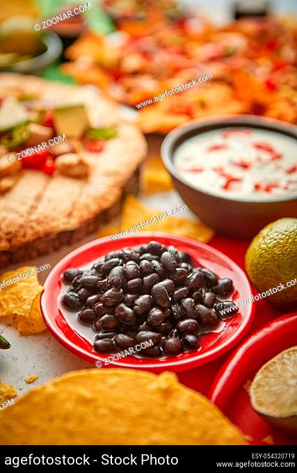 Close up on black beans with various freshly made Mexican foods assortment. Placed on colorful table. With nachos, tacos, tortillas, grilled meat, dips