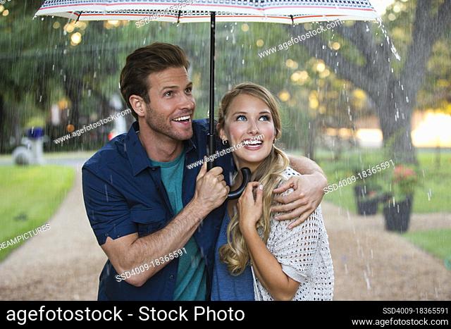 Young couple walking through park with umbrella in the rain, both looking out at the falling rain